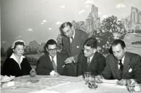 Three men and one women are sitting down and one man is standing up . He is showing something on a paper to others.